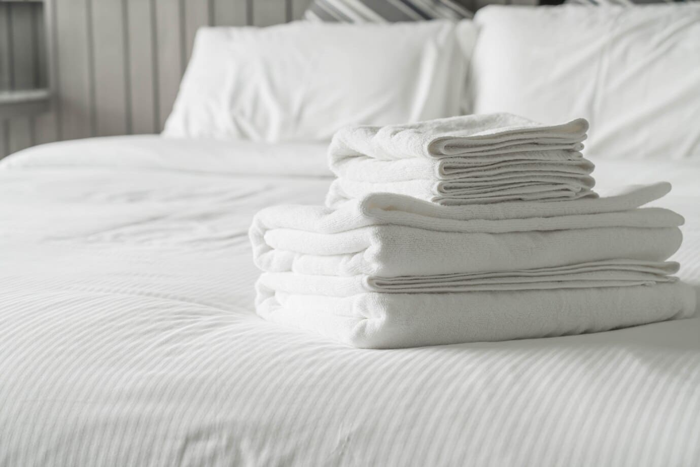 A stack of folded white towels is placed on a neatly made white bed.