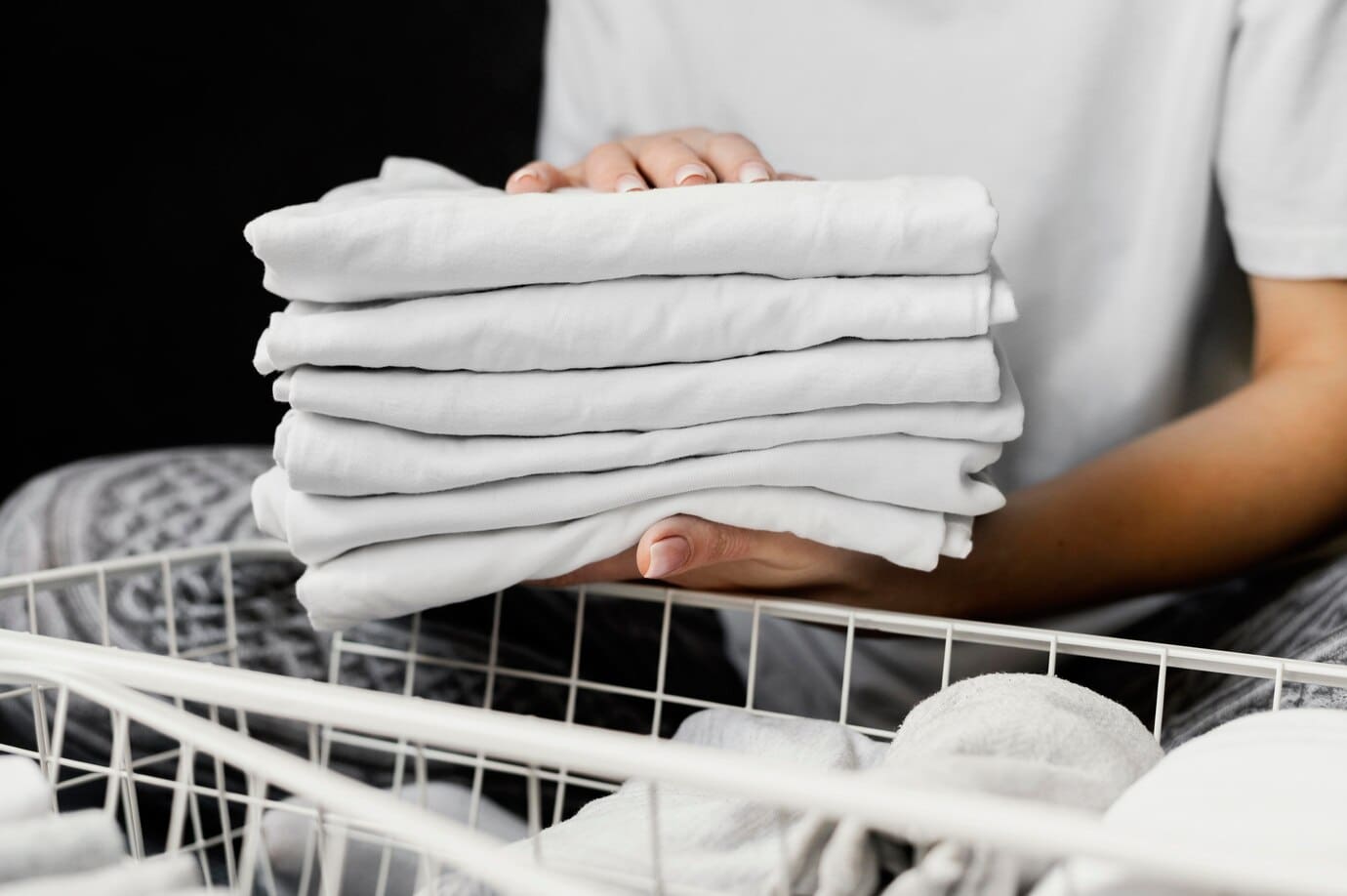 A person holds a neatly folded stack of white shirts above a laundry basket.