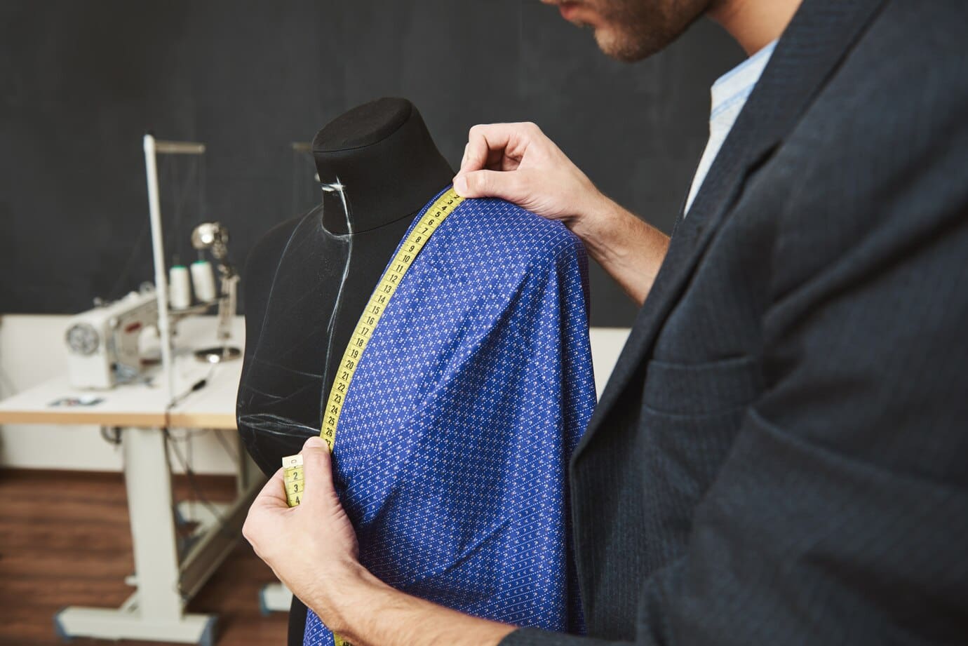 Tailor measuring blue fabric on a dress form with a yellow measuring tape. Sewing machine visible in the background.