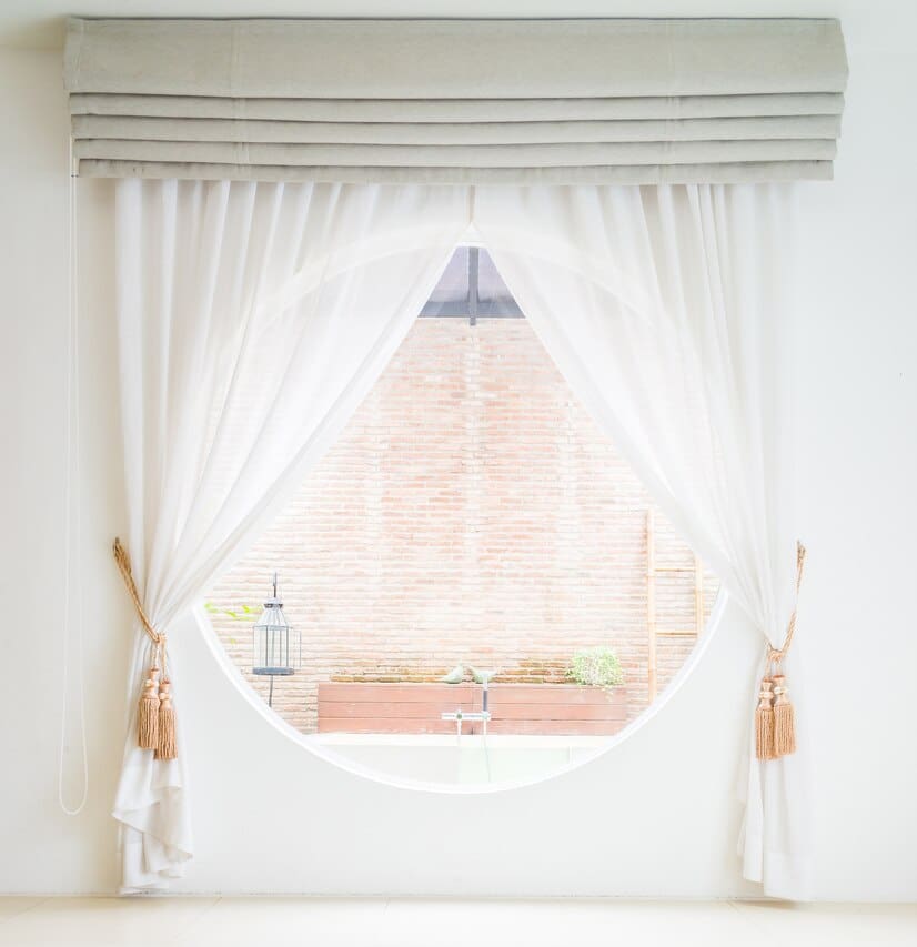 White curtains frame a large circular window, revealing a view of an outdoor space with a brick wall and a small water feature.