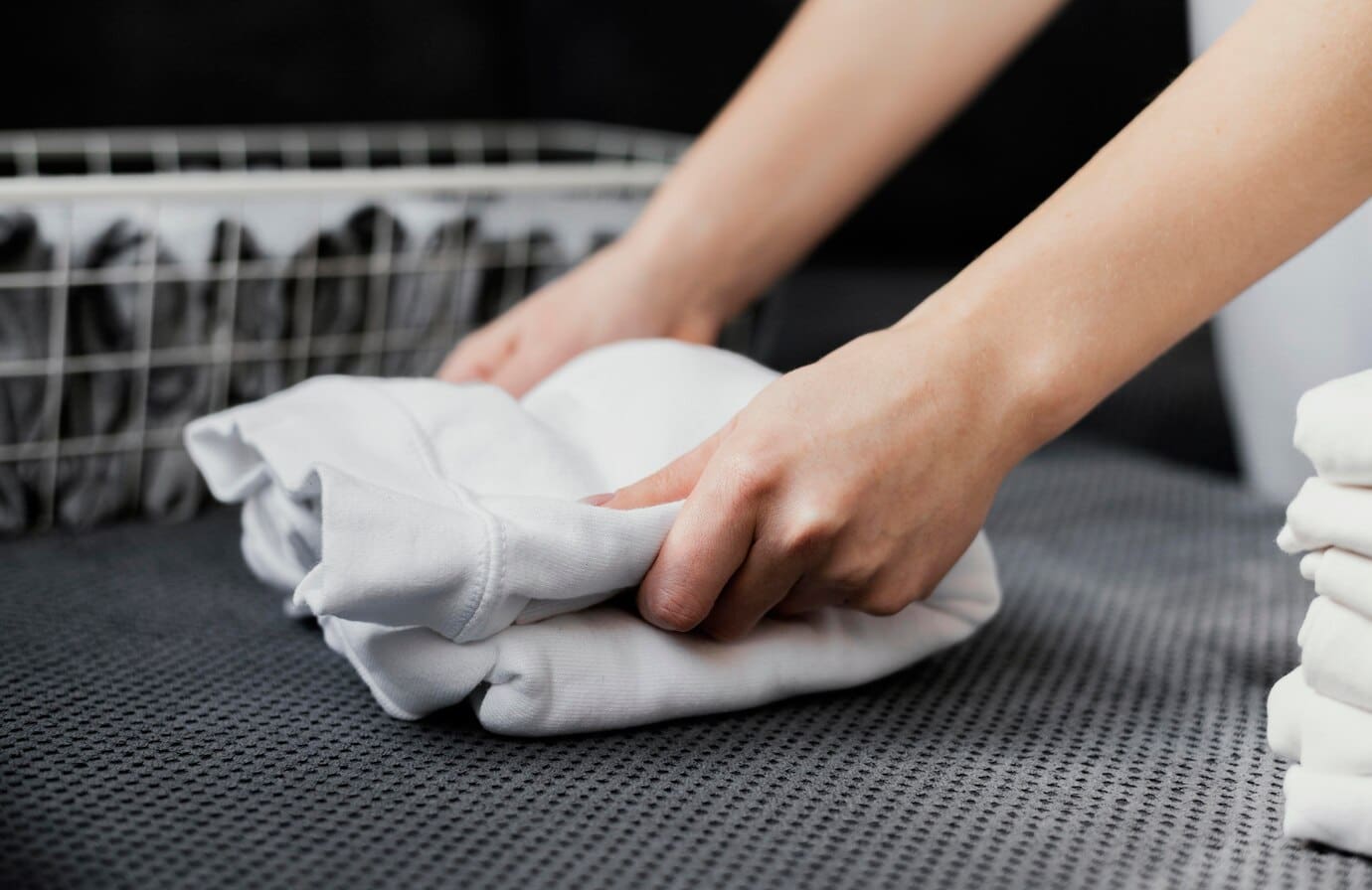 Person folding a white shirt on a gray surface next to a laundry basket with folded clothes.