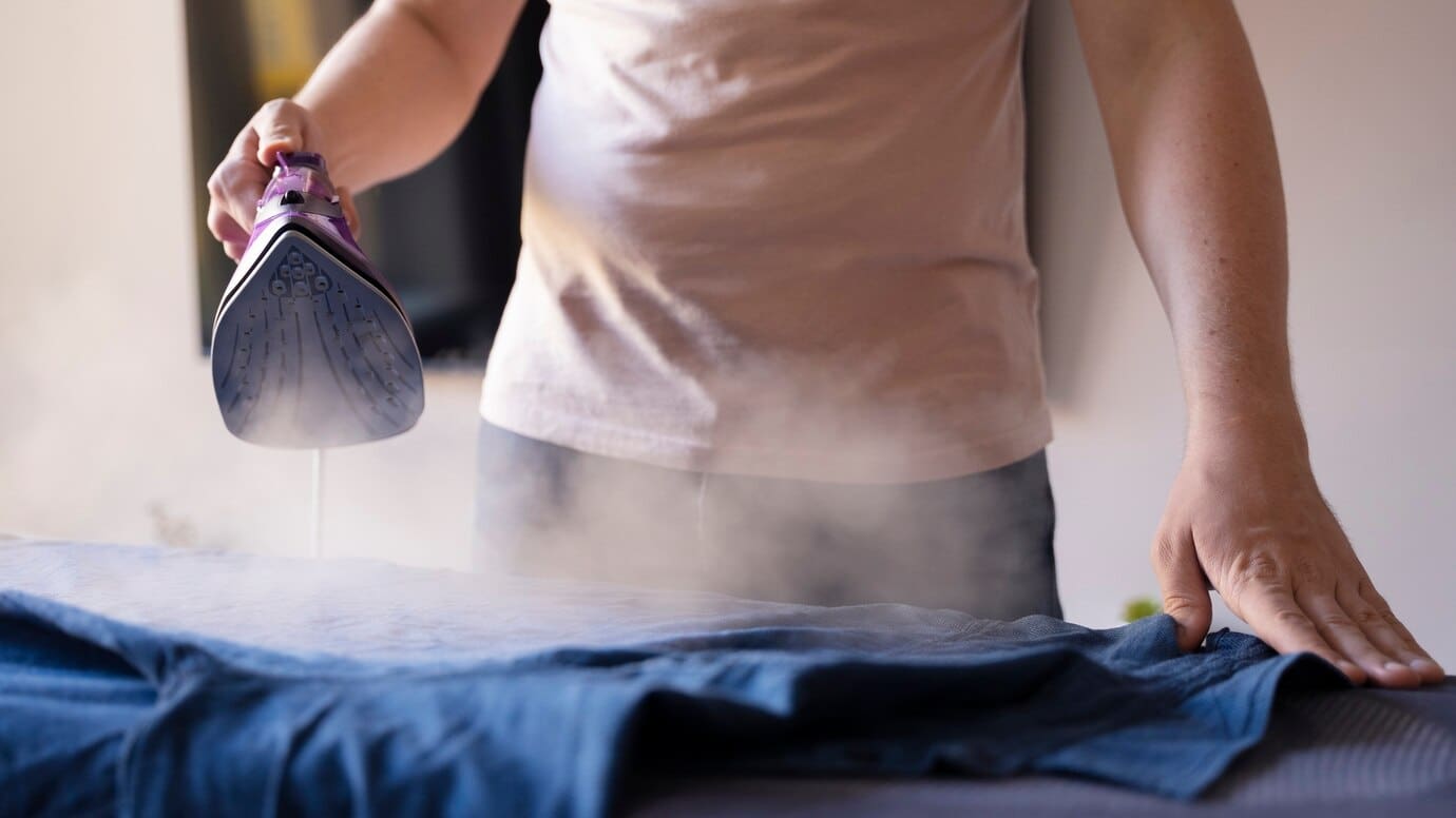 A person is ironing a blue shirt with a purple steam iron, producing visible steam.