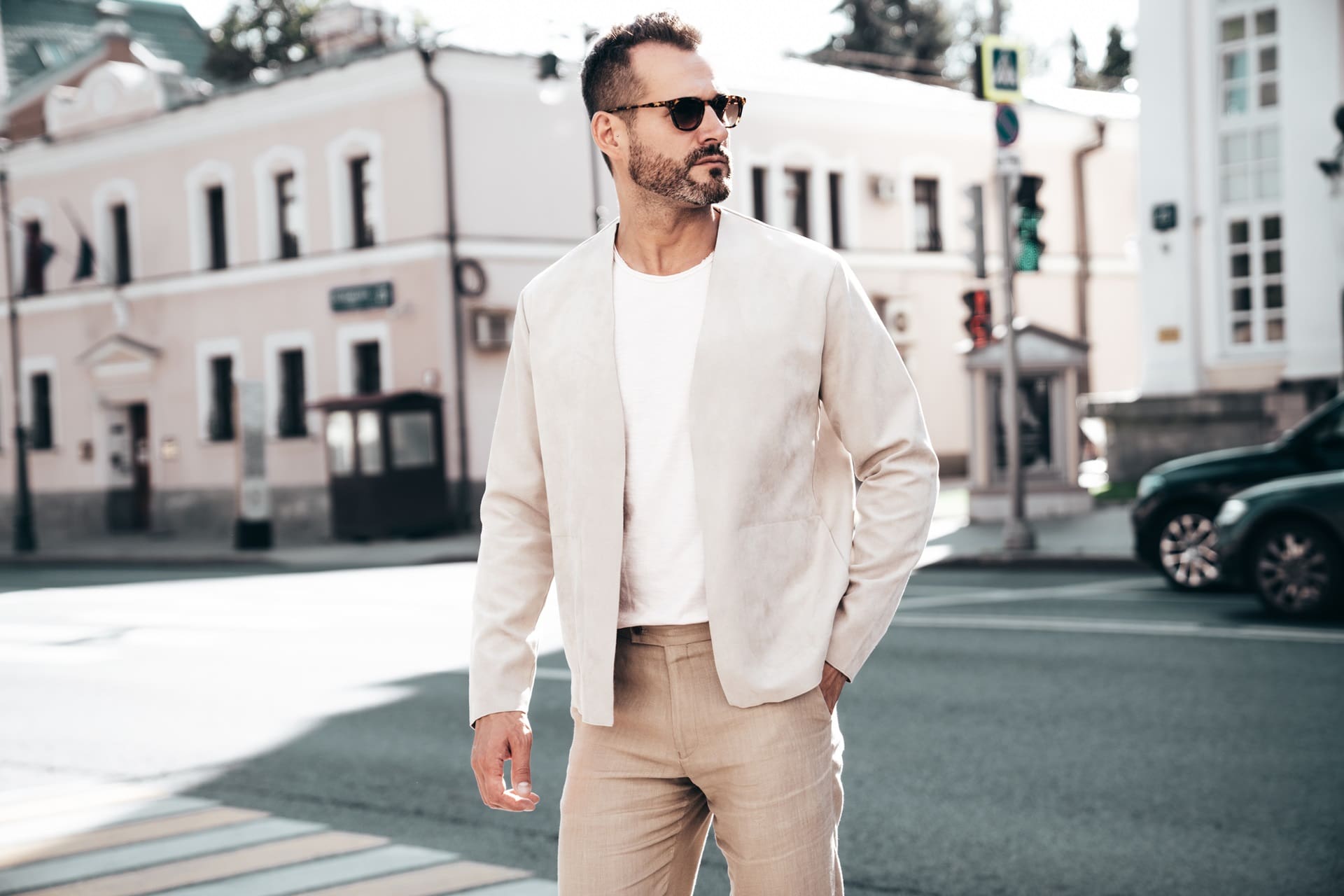 A man wearing sunglasses, a light jacket fresh from dry cleaning, and pants stands confidently on a bustling city street with buildings and cars in the background.