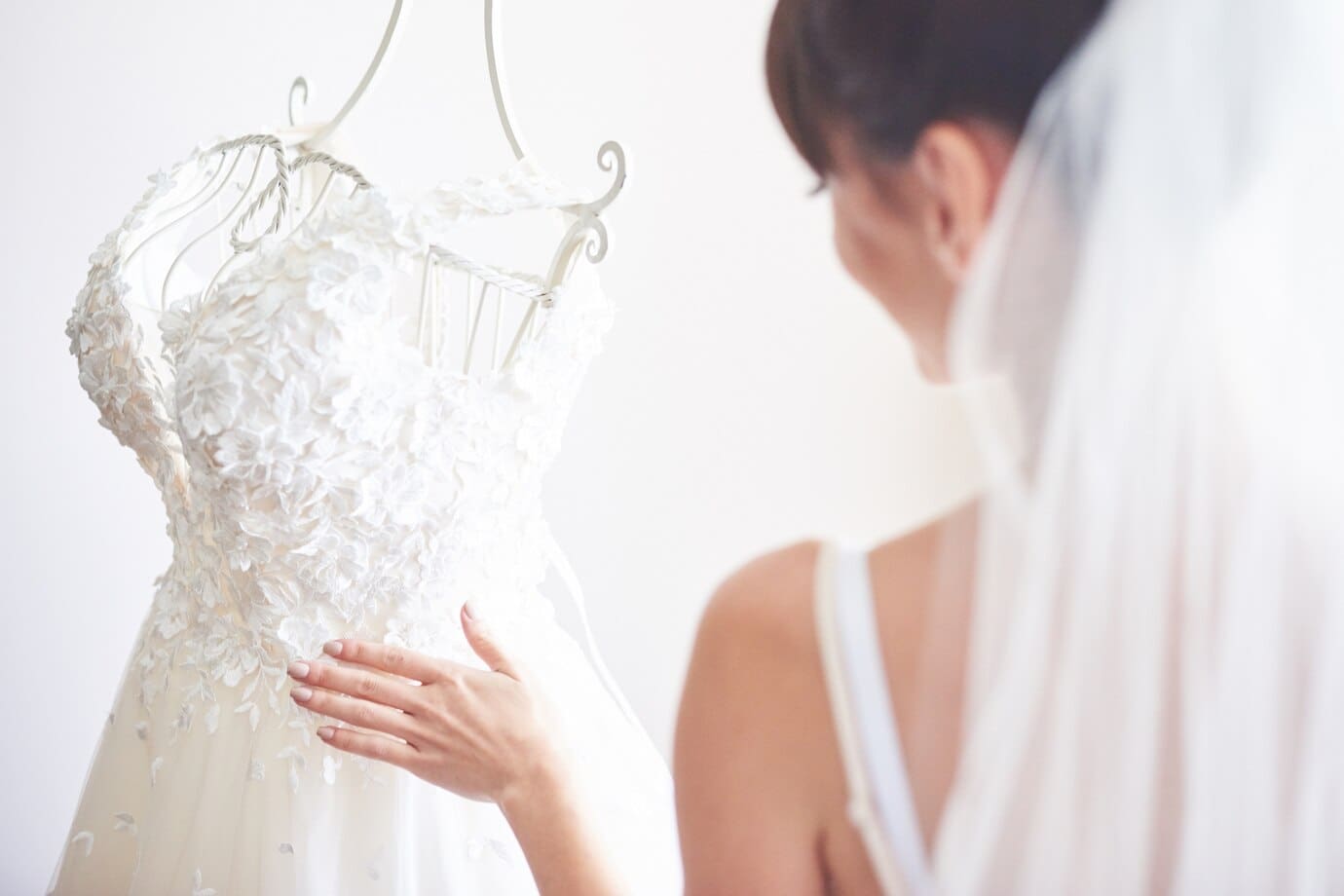 A bride touches a white wedding dress adorned with floral details on a mannequin.