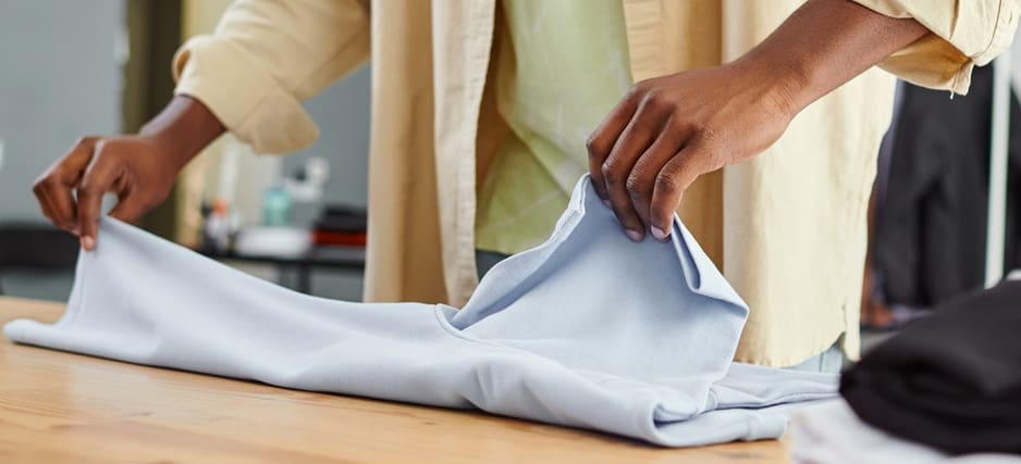 Person folding a light blue garment on a wooden table, wearing a yellow shirt.