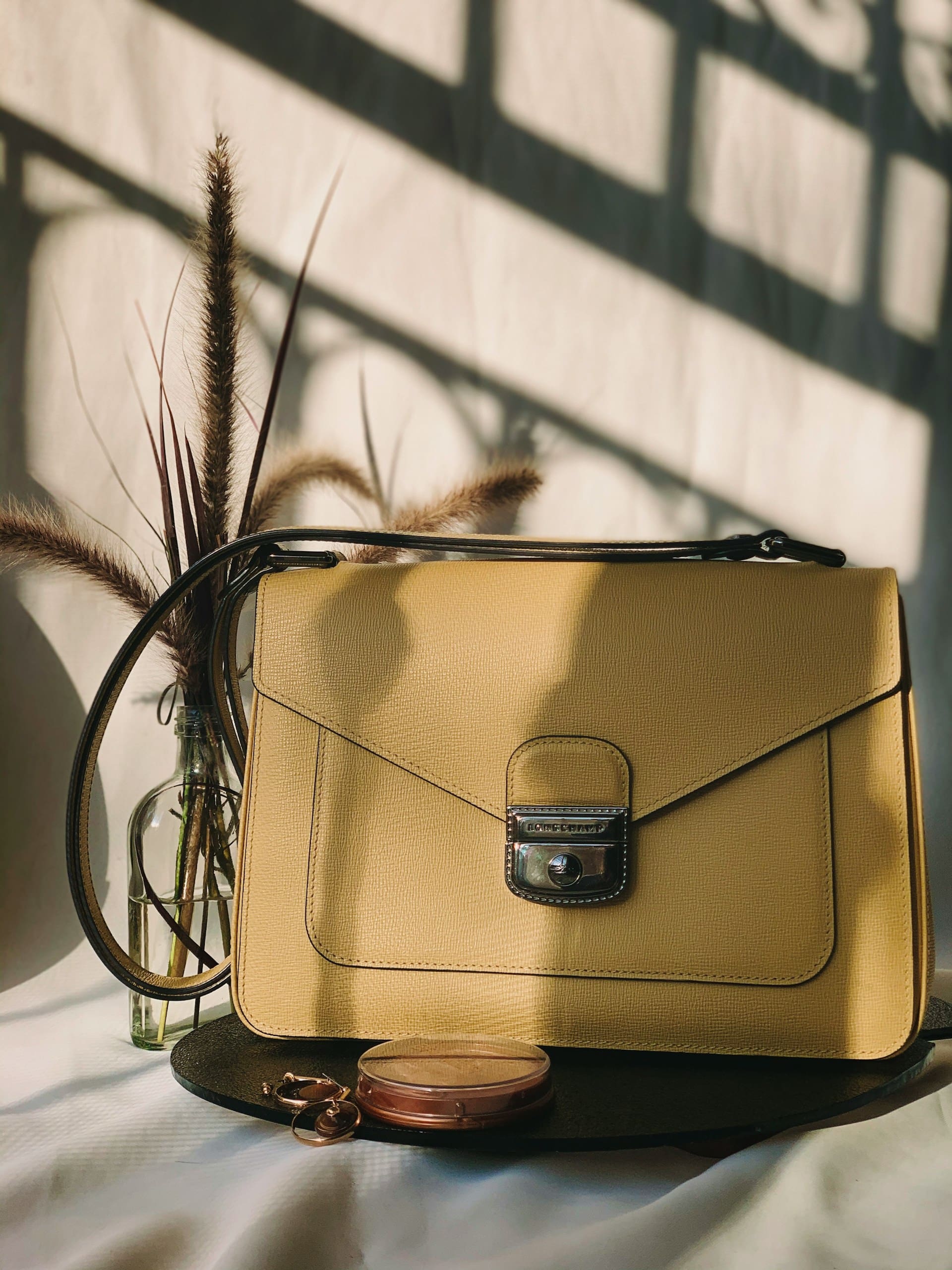 A yellow leather handbag with a front clasp is displayed on a table beside a small glass vase with dried grass and a round coin purse. Sunlight casts shadows in the background.