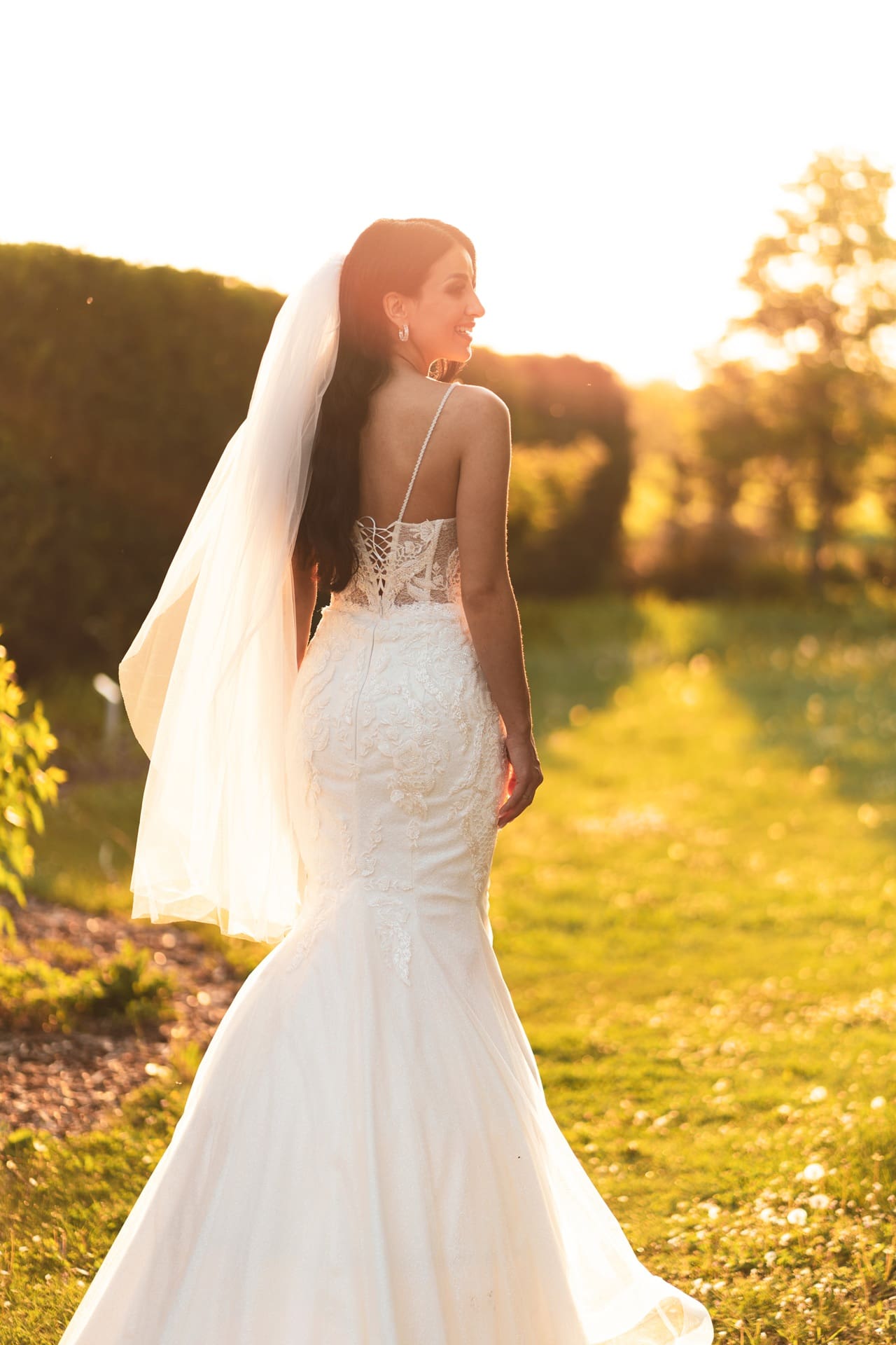 A woman in a white lace wedding dress and veil stands outdoors on a grassy path at sunset, facing away from the camera.
