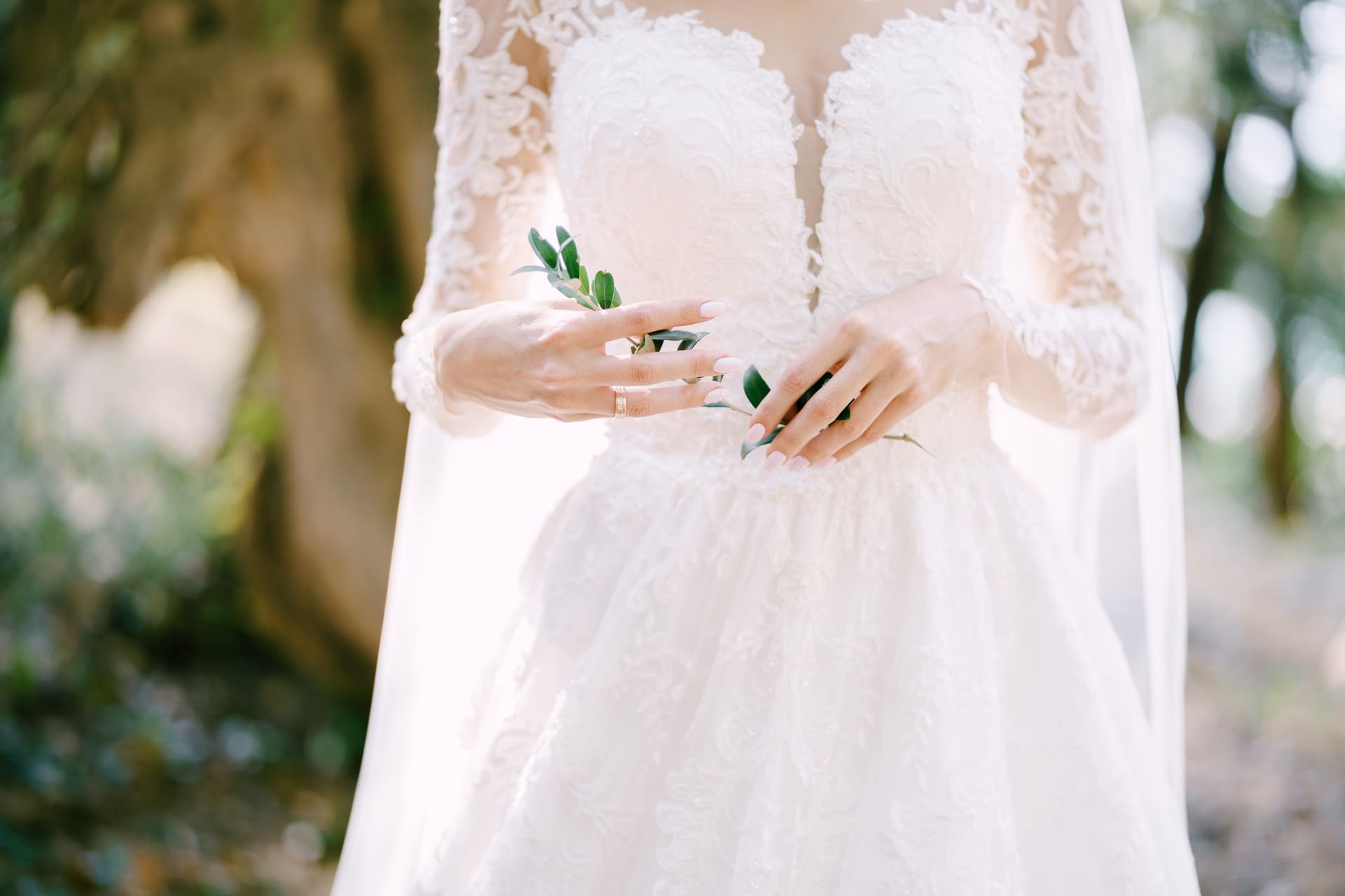 A bride in a white lace wedding dress holds green foliage in her hands, showing her wedding ring. The background is blurred outdoor scenery.
