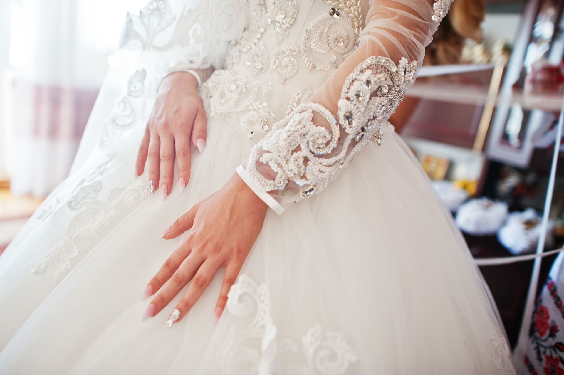 A close-up of a bride’s hands resting on a detailed white wedding dress with lace and bead embellishments.