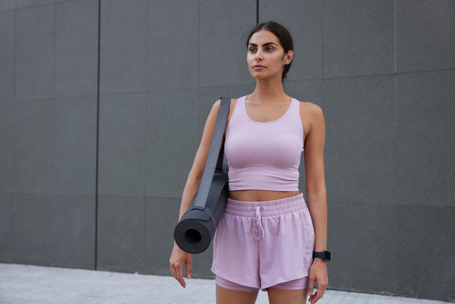 Woman in pink workout clothes holding a rolled yoga mat and wearing a fitness tracker stands outdoors in front of a gray wall.