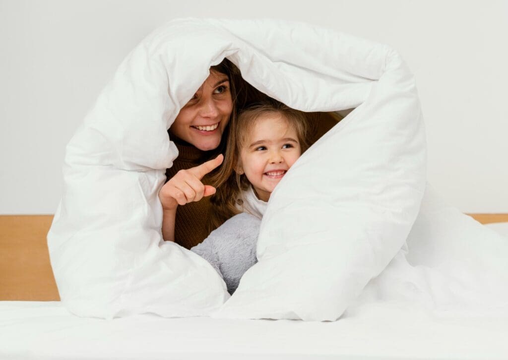 An adult and a child smile and sit under a white blanket on a bed, with the adult pointing at something off-camera.