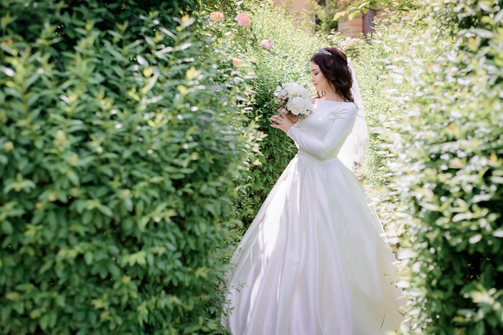 A bride in a white wedding dress holds a bouquet while standing on a path between tall green hedges in an outdoor garden.