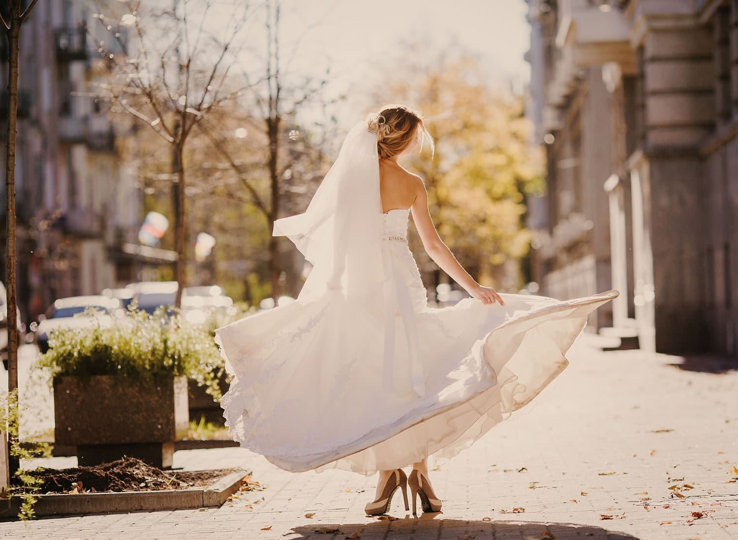 A bride in a white wedding dress and veil twirls on a sunlit city sidewalk, with buildings and trees in the background.