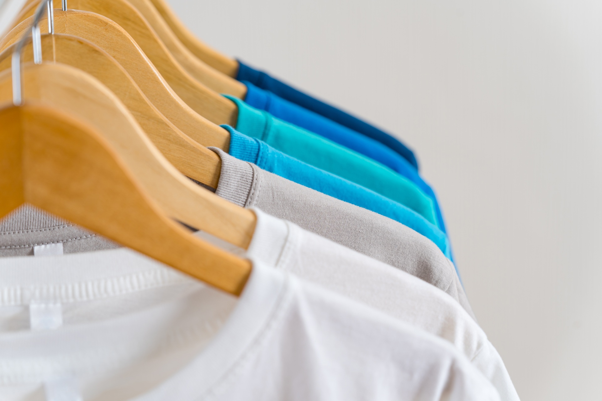 Several t-shirts in shades of white, gray, teal, and blue hang on wooden hangers against a plain background.