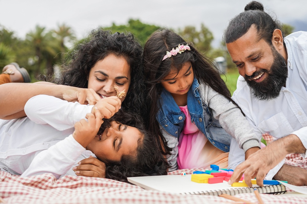 A family of four lays on a picnic blanket outdoors, smiling and playing with colorful blocks and a drawing pad.