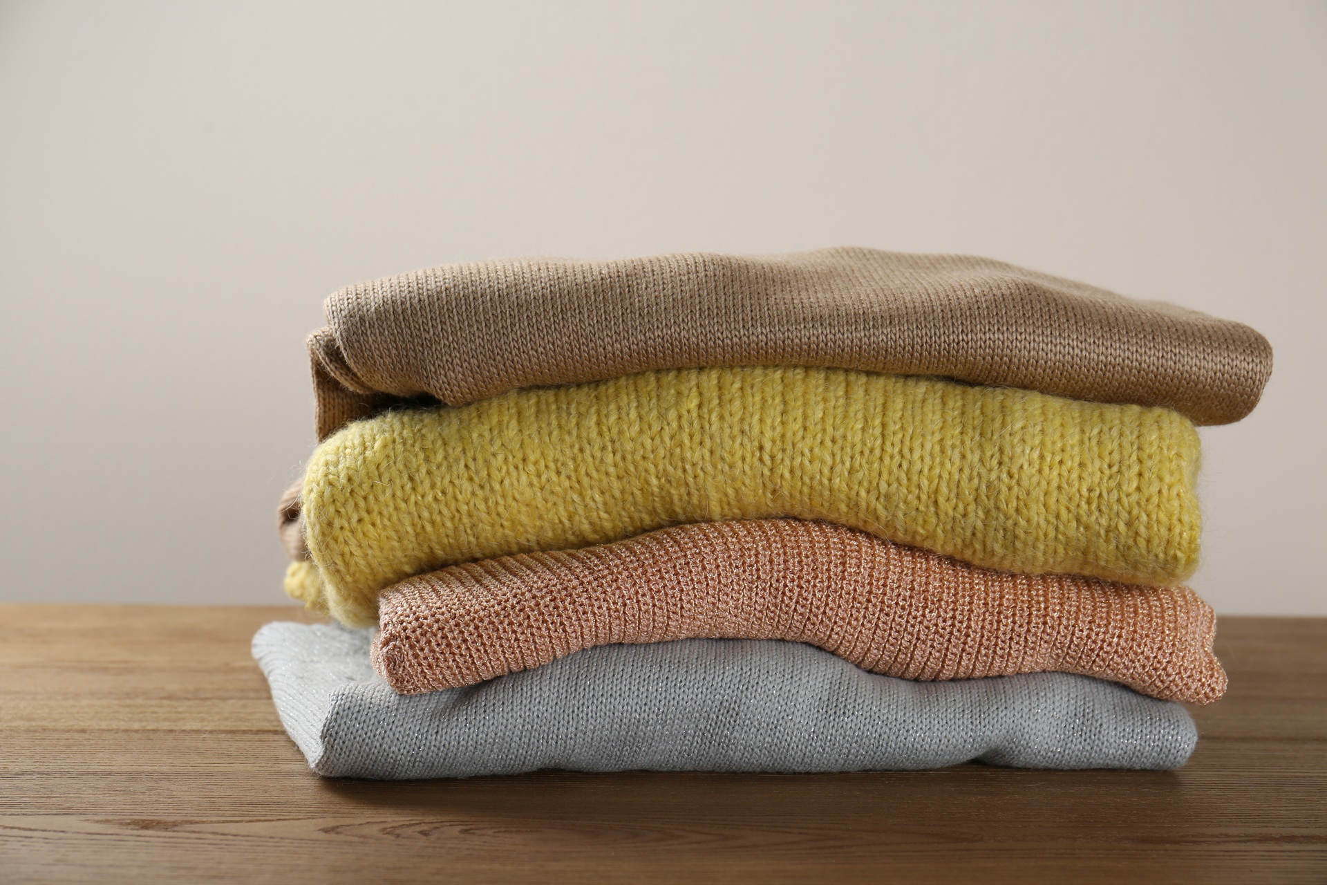 Four folded sweaters in beige, yellow, orange, and gray are stacked on a wooden surface against a neutral background.