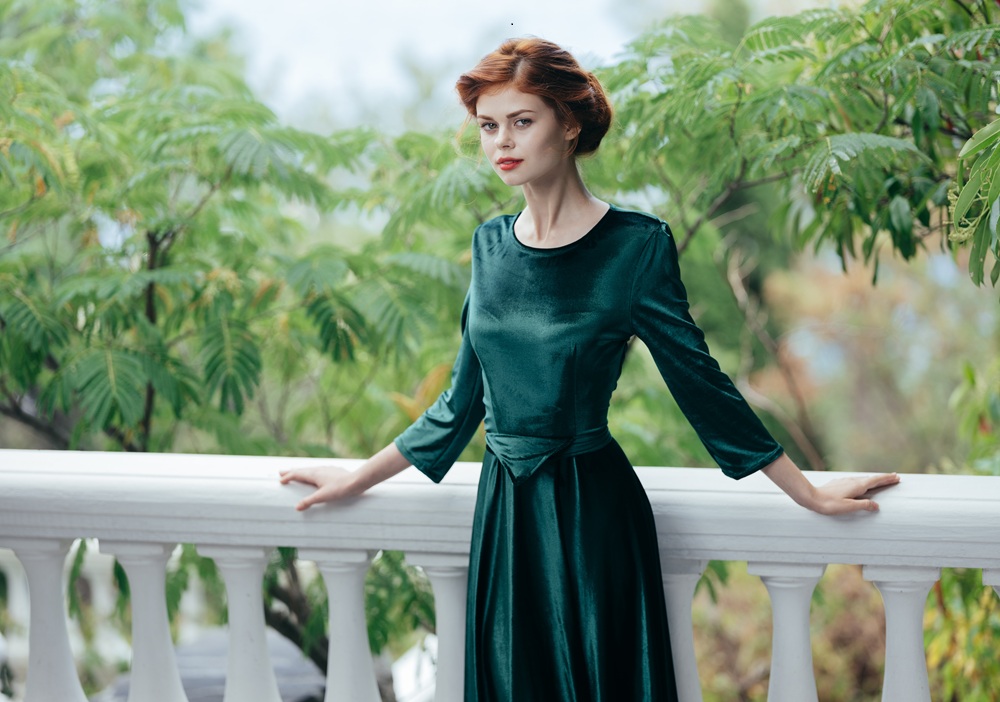 A woman with red hair wearing a long-sleeved green dress stands outdoors, leaning on a white railing with green leafy trees in the background.
