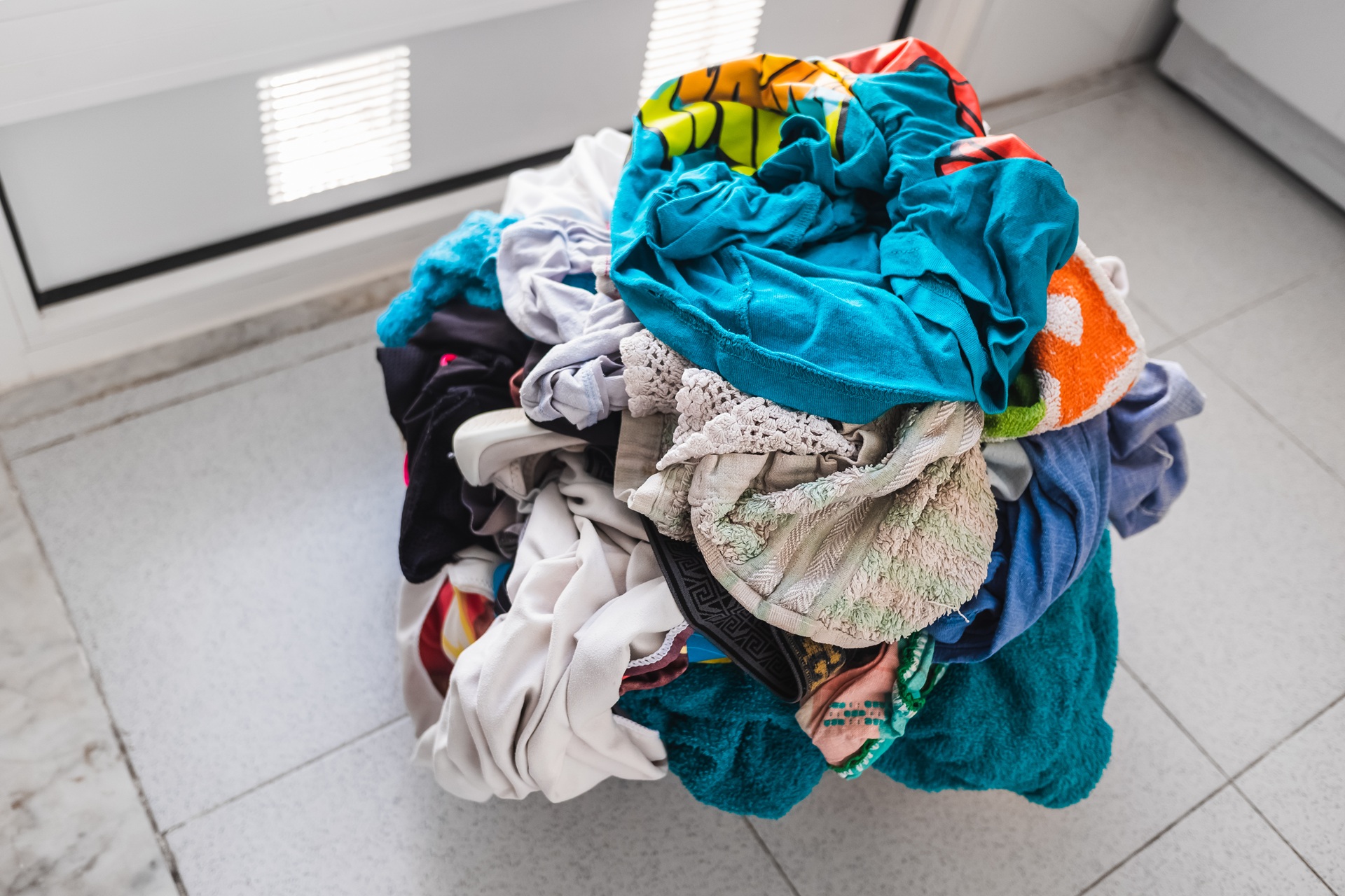 A pile of colorful, mixed laundry clothes sits on a tiled floor near a window with white blinds.