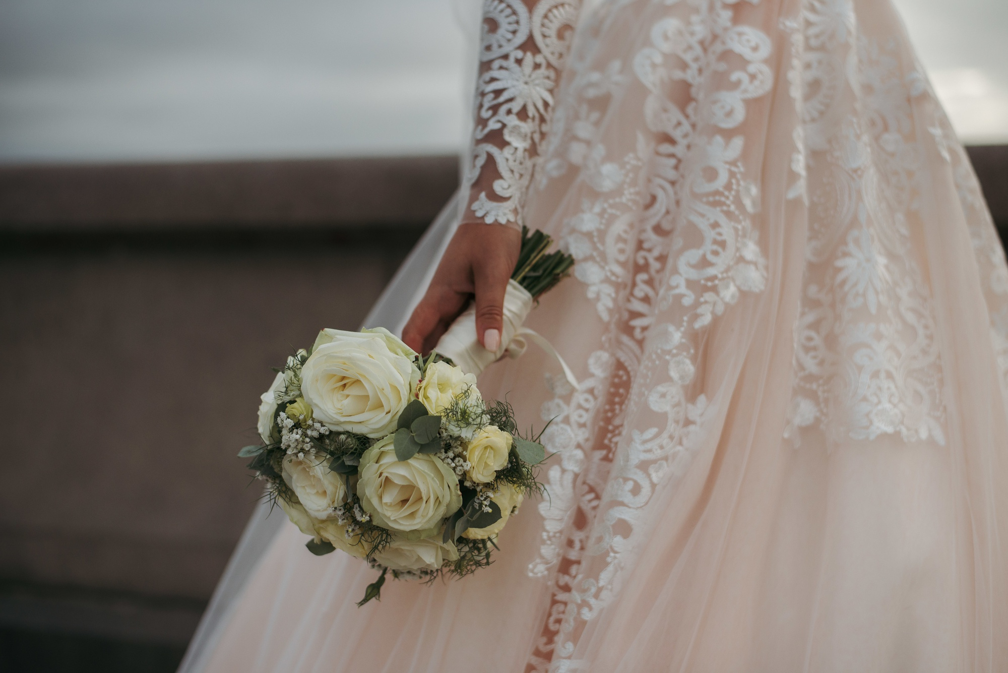 A bride in a lace wedding dress holds a bouquet of white roses and greenery by her side.